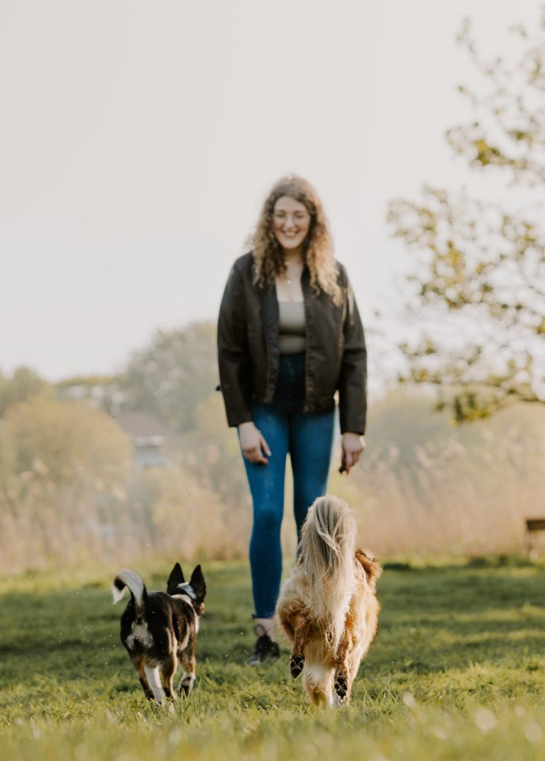 Eine Frau mit lockigem Haar steht auf einer Wiese, zwei Hunde laufen auf sie zu.