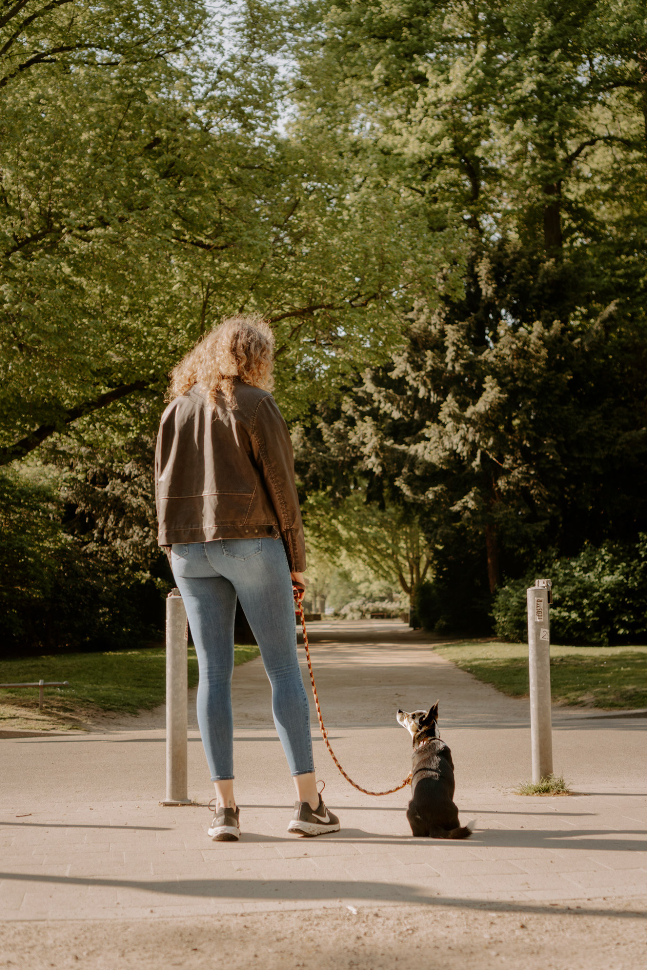 Eine Person mit langen Haaren geht mit einem Hund auf einem Weg im Park.
