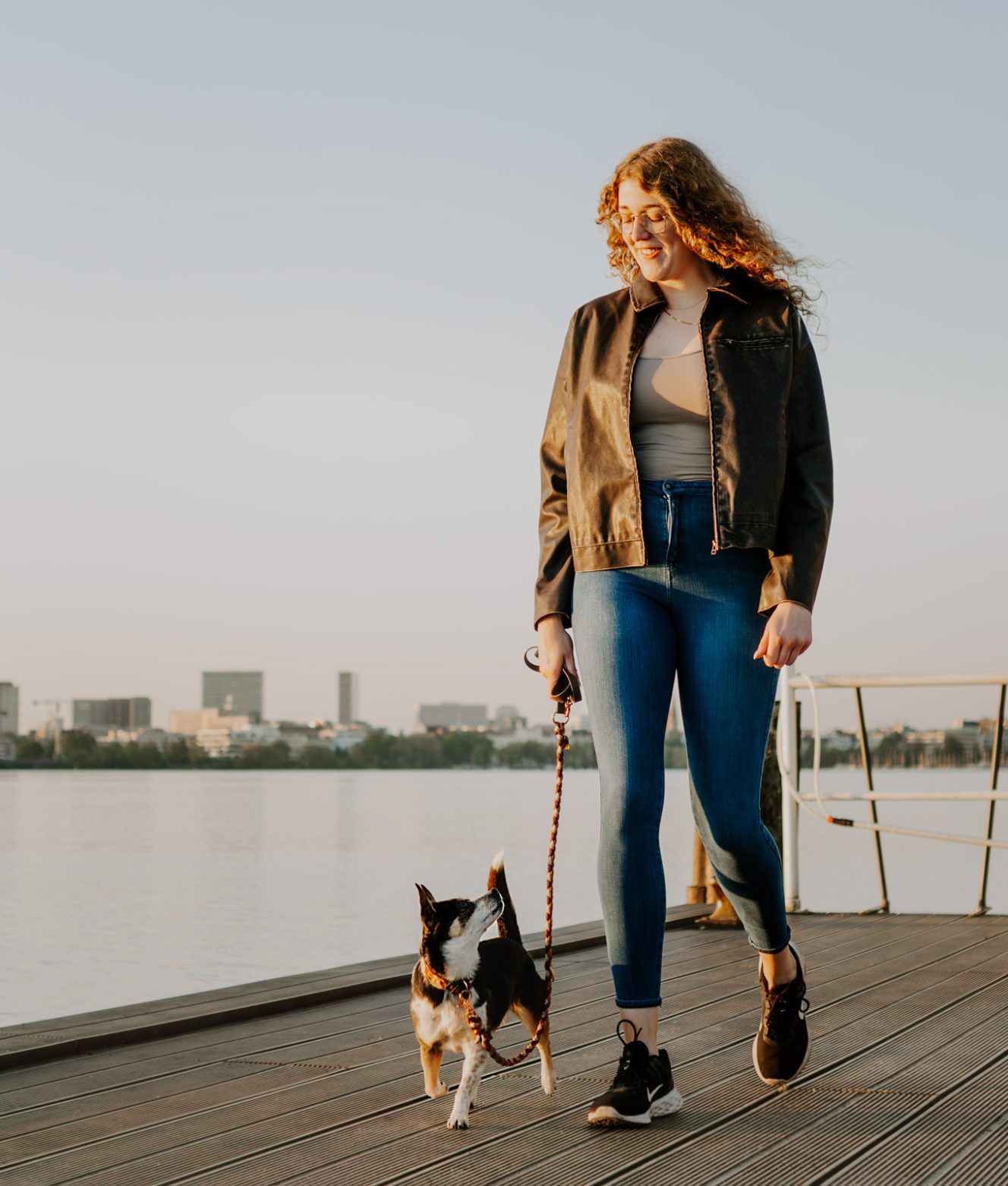 Frau mit blonden Haaren läuft mit einem kleinen Hund am Ufer entlang.