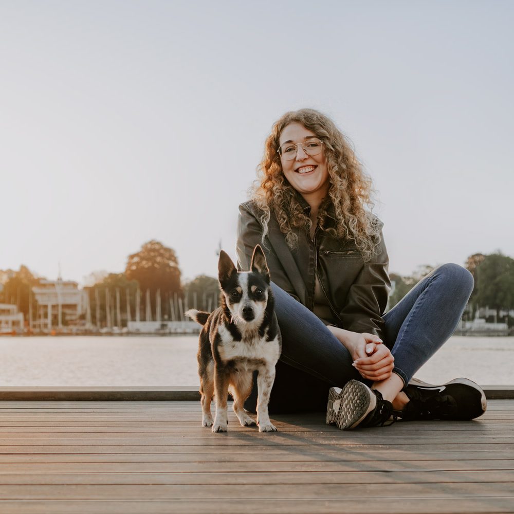 Frau mit lockigem Haar sitzt am Ufer auf einem Steg mit einem Hund.