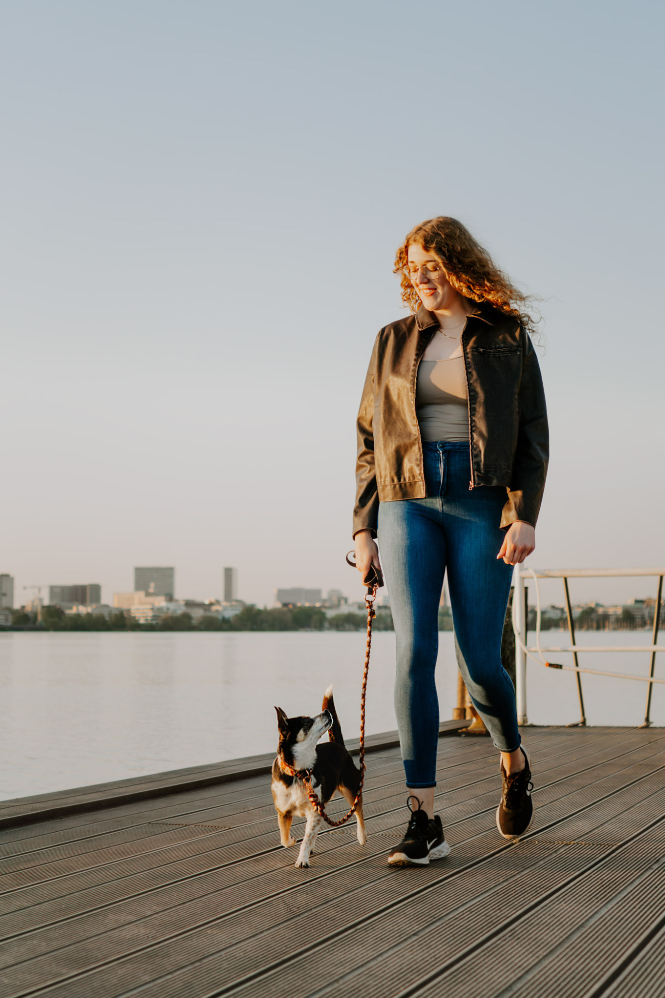 Eine Frau geht mit einem kleinen Hund entlang einer Holzplanke am Wasser.