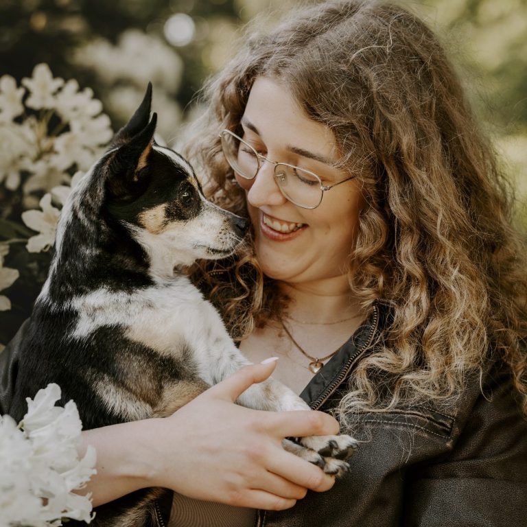 Eine Person mit lockigem Haar hält einen kleinen Hund und lächelt freundlich.